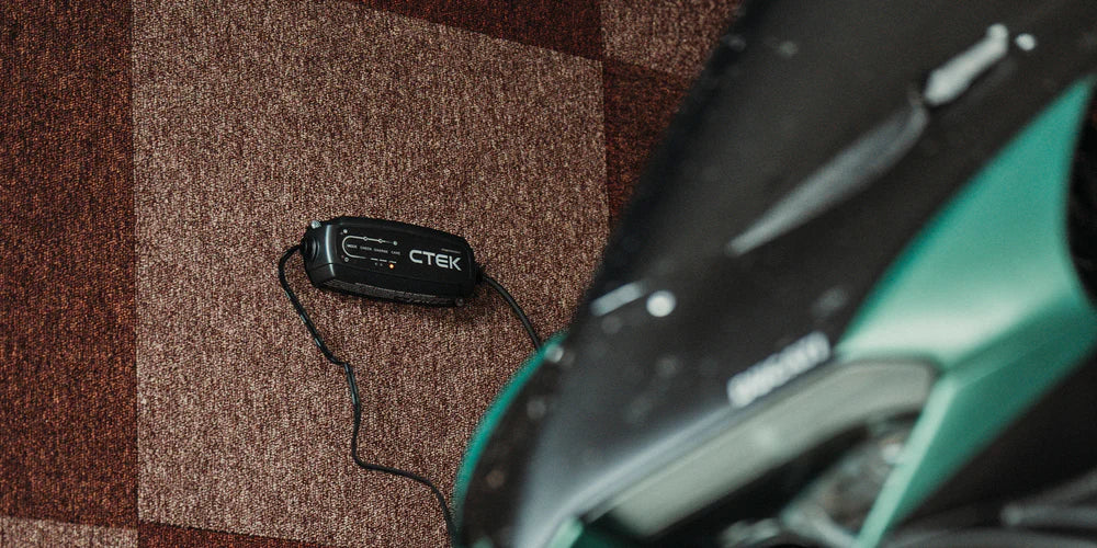 CTEK CT5 Powersport 12V battery charger, black, on a brown carpet beside a green vehicle