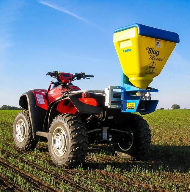 C-Dax SlugMaster SM75 spreader on blue frame mounted to a red ATV in a farm field
