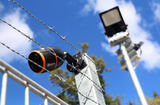 JFC Agri FarmCam Mobility S surveillance camera mounted on a barbed-wire fence; black body with an orange lens ring.