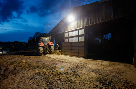 JFC Agri Saber Solar Floodlight mounted on barn at dusk, bright beam over farm yard beside a red tractor.