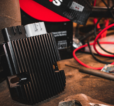 REVOLT regulator rectifier, black finned voltage regulator with gray connector on a workshop bench with red wires