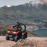 Red ATV quad bike with rider on a dirt trail, rugged mountains and lake in the background.
