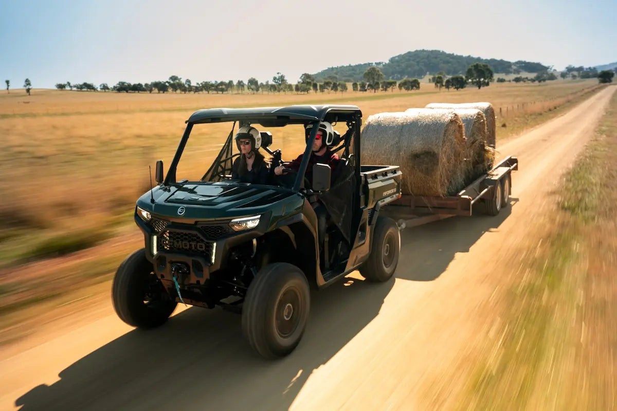 CFMOTO UFORCE U10 Pro Agri UTV in green with two helmeted riders, pulling a trailer loaded with round hay bales.