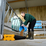JFC Agri Corri feed trough, blue 40L ground feeder in a barn, worker pours feed from a bucket.
