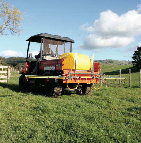 C-Dax spray-on sprayer on ATV with yellow tank and long orange boom arms in a grassy field.