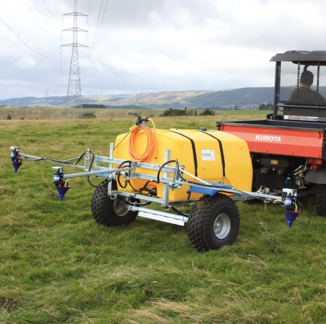 C-Dax Spray-On 500L flat-deck sprayer on trailer hitched to Kubota UTV; yellow tank with blue spray booms in a field.