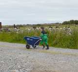 Junior 125L tipping wheelbarrow blue by JFC Agri, puncture-proof wheels, being pushed by a child along a gravel path